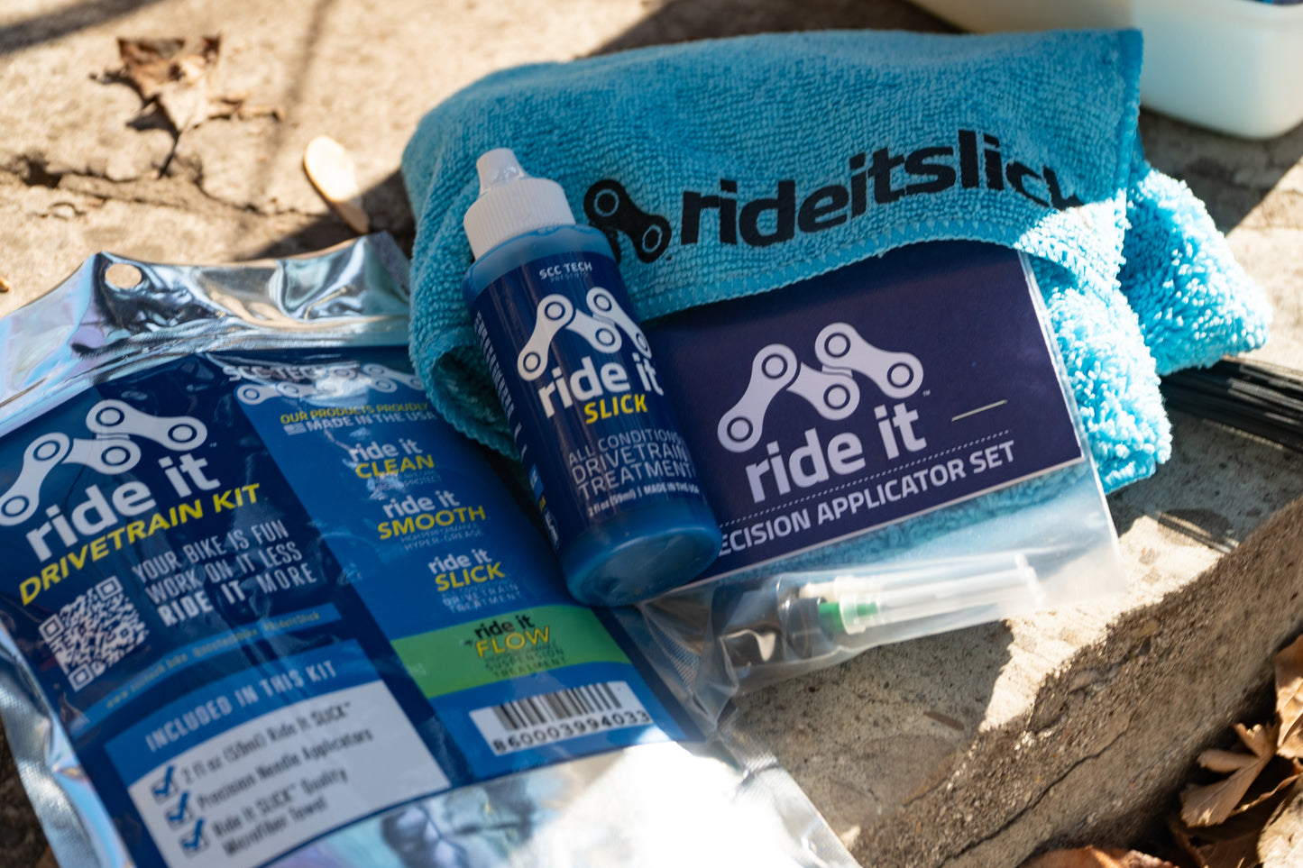 Ride It Drivetrain Kit by SCC Tech, featuring a complete bike maintenance solution. The packaging displays the Ride It brand logo and includes products like Ride It Slick, Ride It Clean, Ride It Smooth, and Ride It Flow. The kit contains 2 fl oz (59 ml) bottle of Ride It Slick drivetrain treatment, precision needle applicators, and a microfiber towel. The label highlights the kit’s focus on enhancing bike performance and protection, with bold text encouraging less time working on it and more time riding it