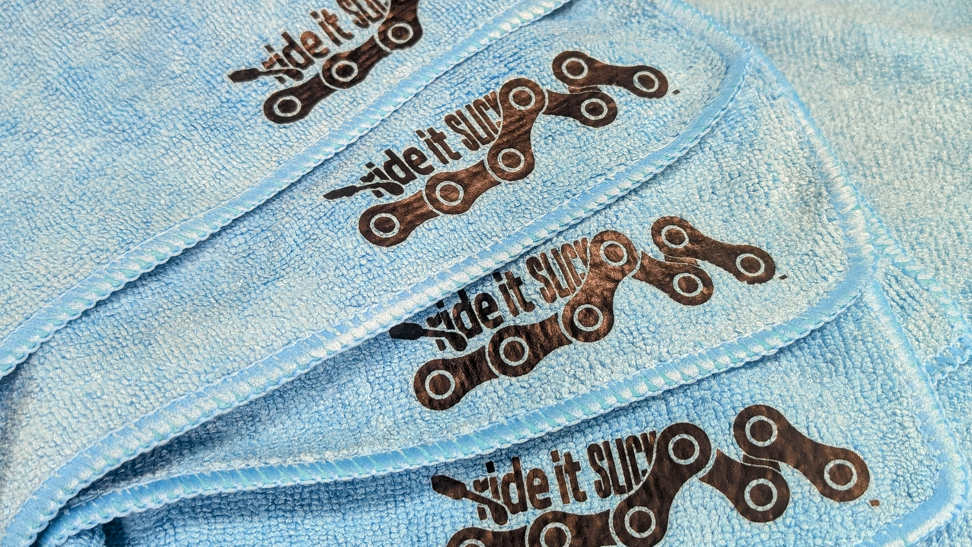 Blue microfiber towel with a printed black bicycle chain logo, designed for cleaning and maintaining bicycles. The soft, high-quality microfiber material is ideal for wiping down bike frames, components, and drivetrains, helping to remove dirt, grime, and excess lubricant during maintenance. This towel is part of the Ride It bike care kit, offering a gentle yet effective cleaning solution.