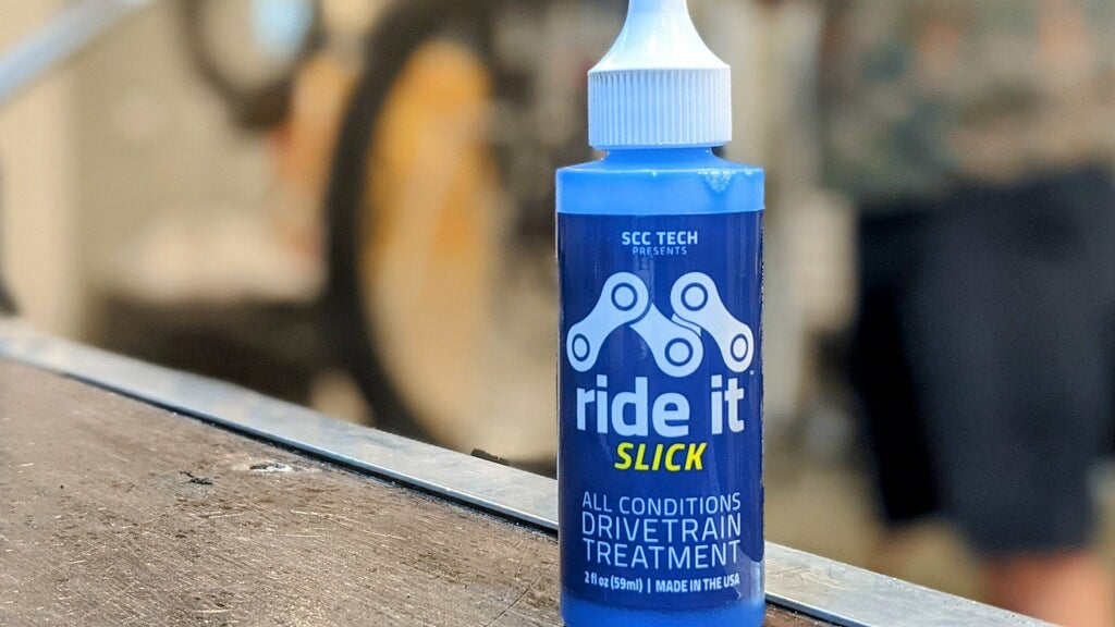 Blue bottle of Ride It Slick all-conditions drivetrain treatment for bicycles, presented by SCC Tech. The 2 fl oz (59 ml) bottle has a white cap and a blue label with bold white and yellow text. The label features a bicycle chain link graphic, highlighting its function as a drivetrain treatment designed to improve performance in all weather conditions. Made in the USA